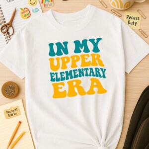 Op de afbeelding: Wit T-shirt met de tekst "IN MY UPPER ELEMENTARY ERA" in turquoise en gele retro lettertype. Het shirt is in de taille geknoopt. Schoolspullen, waaronder een bril, schaar en potloden, zijn rond het shirt geplaatst.