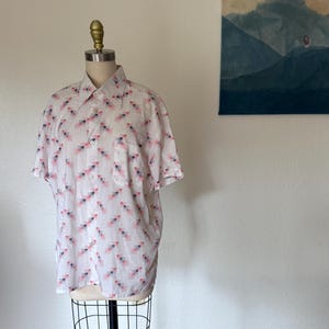 以下が含まれることがあります： マネキンに着せられた半袖の白いシャツ。 ピンクと青の幾何学模様が繰り返されています。 胸ポケットとクラシックな襟が付いています。