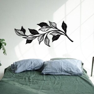 May include: A black metal wall decor featuring a stylized branch with leaves. The decor is mounted on a white wall above a green bed with blue pillows.
