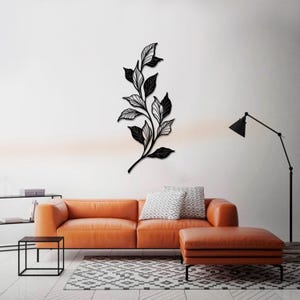 May include: Black metal wall art featuring a stylized branch with leaves. The art is displayed on a white wall above a brown leather couch and a black end table.