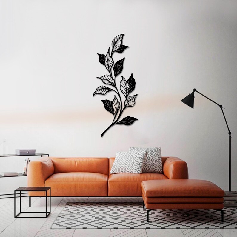 May include: Black metal wall art featuring a stylized branch with leaves. The art is displayed on a white wall above a brown leather couch and a black end table.