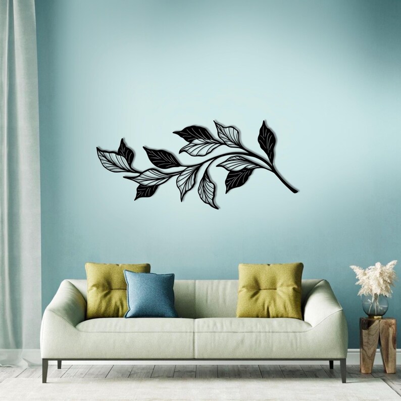 May include: Black metal wall art featuring a stylized branch with leaves. The art is displayed on a light blue wall above a white couch with yellow and blue throw pillows.