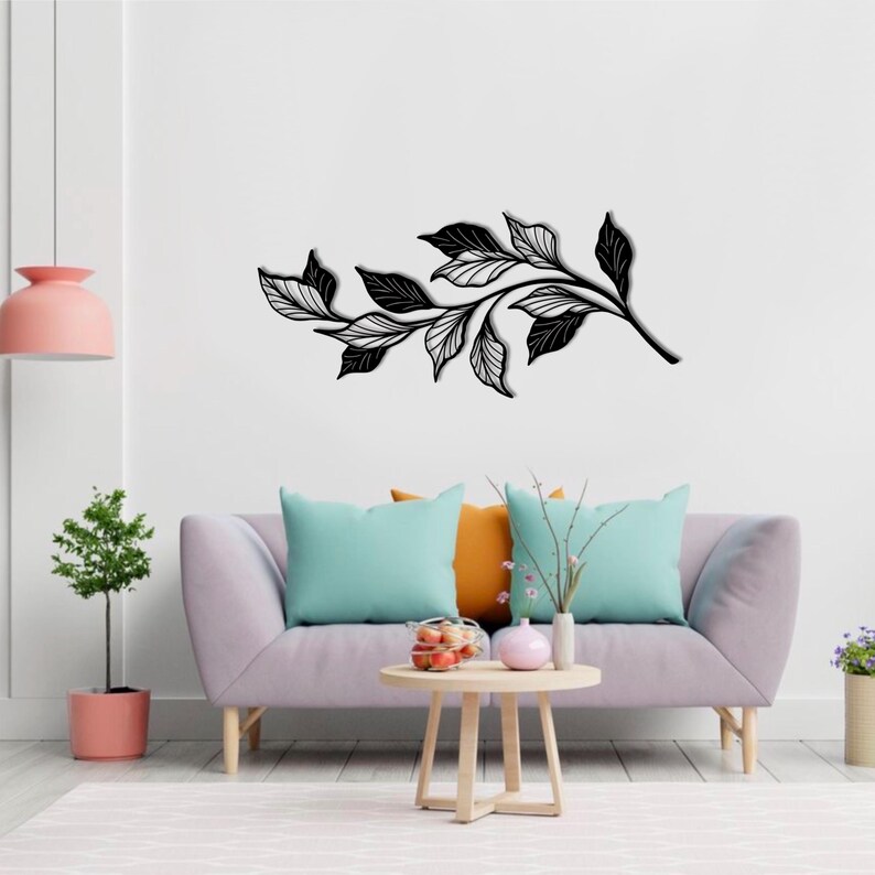 May include: Black metal wall art featuring a stylized branch with leaves. The art is displayed on a white wall above a light purple sofa with teal and orange throw pillows.