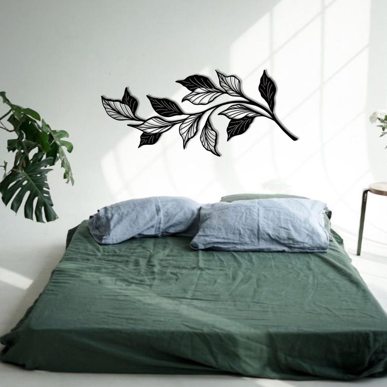 May include: A black metal wall decor featuring a stylized branch with leaves. The decor is mounted on a white wall above a green bed with blue pillows.