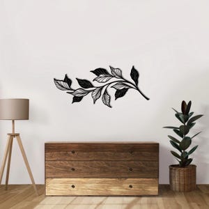 May include: Black metal wall art featuring a stylized branch with leaves. The branch is curved and has a delicate, intricate design. The leaves are arranged in a flowing pattern.