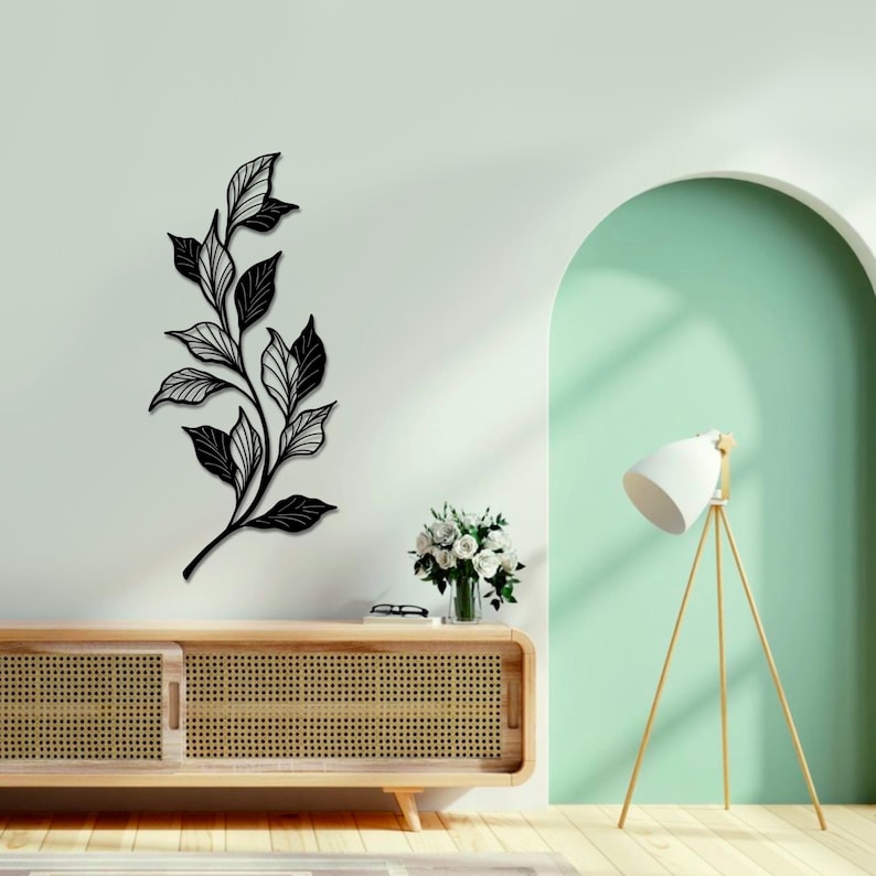 May include: Black metal wall art featuring a stylized branch with leaves. The art is mounted on a white wall above a wooden cabinet with a woven rattan front. A white lamp with a tripod stand is in the background.