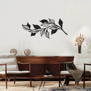 May include: Black metal wall art featuring a stylized branch with leaves. The art is displayed on a white wall above a wooden cabinet and two chairs.