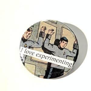 May include: A circular pinback button featuring a vintage comic book illustration of two figures in grey uniforms with raised arms. A white rectangular strip is overlaid across the center with the black printed text, I love experimenting.
