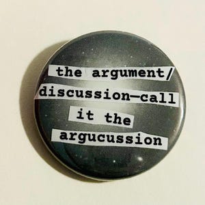 May include: A round button with a dark gray and white speckled background. The text "the argument/ discussion-call it the argucussion" is printed on the button in a typewriter font.