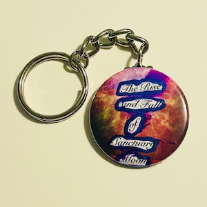 May include: A round keychain with a silver chain and ring. The circular pendant features a colourful design with the text "The Rise and Fall of Sanctuary Moon" in blue and white lettering against a red and purple background.