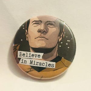 May include: Round button with a portrait of a man and the words "Believe in Miracles" on a white background. The man's portrait is in shades of brown, with a dark background and white star-like dots.