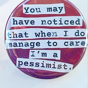 May include: A circular, maroon badge with the quote "You may have noticed that when I do manage to care I'm a pessimist." The text is in a typewriter font on white rectangles.