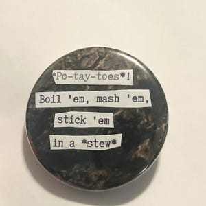 May include: A round, dark-colored button with the text "*Po-tay-toes*! Boil 'em, mash 'em, stick 'em in a *stew*". The button has a textured, mottled appearance, with a glossy finish. The text is in a typewriter-style font.