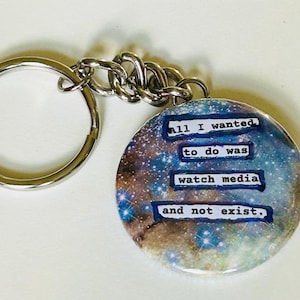 May include: A round keychain featuring a galaxy design and the text "all I wanted to do was watch media and not exist." It has a silver keyring and chain.