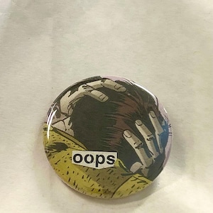 May include: A round button with a comic book illustration of a person holding their head in their hands. The word "oops" is printed in a white, sans-serif font on a yellow background.