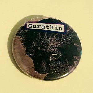 May include: A circular badge featuring a dark, textured design and the word "Gurathin" in white text on a rectangular label. The badge has a dark background with hints of blue and purple.