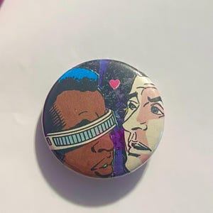 May include: Circular button with a comic book illustration. It depicts two faces, one with a blue headband and the other with a surprised expression. A pink heart is above the faces. The background is purple and black.