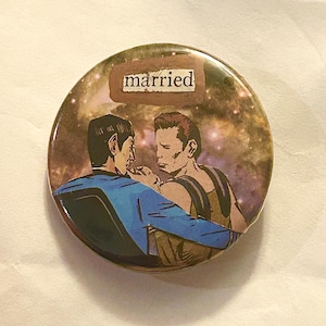 May include: Round button with a galaxy background and an illustration of two men embracing. The word "married" is printed on a brown rectangle at the top of the button. The men are in a romantic pose.