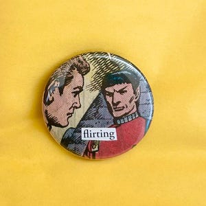 May include: A round button with a comic book style illustration of two men. One man is in profile, the other is in a red uniform. The word "flirting" is printed in a white rectangle.