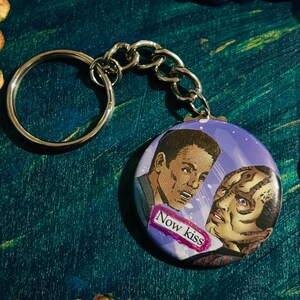 May include: A silver keychain with a round button featuring a comic book style image of two men. The button has a purple background with white stars and the text "Now kiss" in pink.