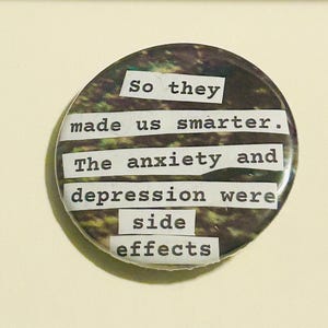 May include: A circular badge featuring a forest-themed design and the text "So they made us smarter. The anxiety and depression were side effects" in a typewriter font. The badge has a dark, glossy finish.