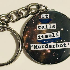 May include: A circular keyring with a silver chain and ring. The keyring features a galaxy-themed design and the text "It calls itself 'Murderbot'" in white block letters.