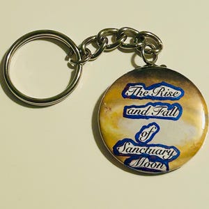 May include: A circular keyring with a silver chain and ring. The pendant has a celestial design and the text "The Rise and Fall of Sanctuary Moon" in blue and white.