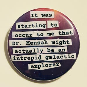 May include: A circular badge with a purple and red galaxy design. The text on the badge reads: "It was starting to occur to me that Dr. Mensah might actually be an intrepid galactic explorer."