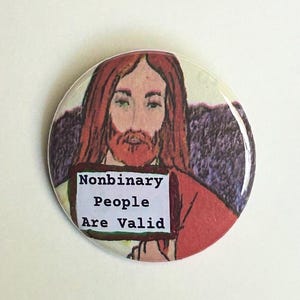 May include: A round button with an illustration of a person with long brown hair and a beard, wearing a red robe. The button has the text "Nonbinary People Are Valid" on a white background.