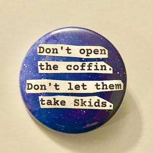May include: A circular badge with a dark blue and purple galaxy pattern. The text "Don't open the coffin. Don't let them take Skids." is printed in white, typewriter-style font.