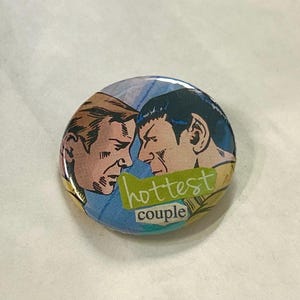 May include: Round button featuring a comic-style illustration of two men facing each other. The button has a blue and yellow background with the words "hottest couple" in green and white.
