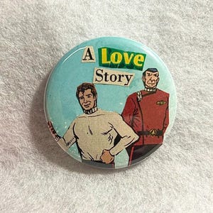 May include: A round button with a light blue background featuring two illustrated figures and the text "A Love Story." One figure wears a white turtleneck, and the other wears a red uniform. The text "Love" is in yellow.