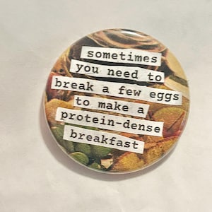 May include: A circular button with a colourful background. The text on the button reads, "sometimes you need to break a few eggs to make a protein-dense breakfast." The button has a glossy finish.