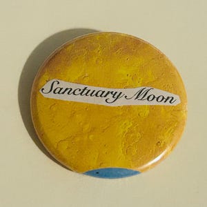 May include: A circular badge with a textured yellow surface and the words "Sanctuary Moon" on a white banner. A small blue arc is at the bottom. The badge has a glossy finish and a shadow.