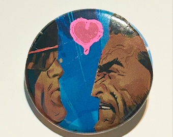 May include: A round button with a blue background and two side profiles of men facing each other. A pink heart shape is above the men. The button has a silver metal edge.