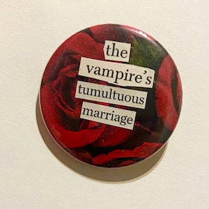 May include: A round button with a red rose background and the text "the vampire's tumultuous marriage" in white block letters. The button has a glossy finish and a slight shadow.