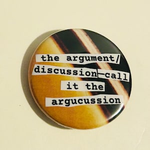 May include: A circular badge with a brown and black striped design. The text "the argument/ discussion-call it the argucussion" is printed on the badge in a typewriter font. The badge has a glossy finish.