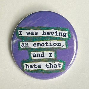May include: A circular purple badge with the words "I was having an emotion, and I hate that" in white text with green glitter edging. A simple, expressive accessory.