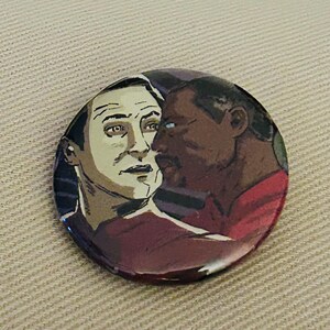 May include: A round button featuring a close-up portrait of two men. The man on the left has pale skin and dark hair. The man on the right has dark skin and dark hair. Both men are looking at each other. The button has a red, white, and blue color scheme.