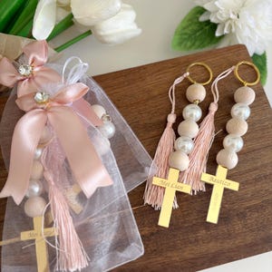May include: Two keyrings with wooden beads, faux pearls, and gold crosses. One keyring is in a sheer bag with a pink bow. The crosses are engraved with names and the word "Baptizo". White flowers are in the background.
