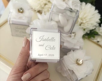 May include: Clear square favour boxes filled with white sweets, decorated with white fabric flowers and a gold gem centre. Each box has a silver label that reads "Isabelle and Cole, April 17, 2026".