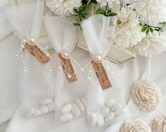 May include: Three small, clear mesh bags filled with white candies, tied with tulle and adorned with pearl accents and tags that read "Our Wedding". White flowers and decorative flowers are in the background.
