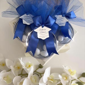 Pode incluir: Três caixas de presente com cestos de arame prateado cheios de doces brancos, adornadas com tule azul, laços e etiquetas que dizem "Dottoressa Natascia". Flores de orquídeas brancas em primeiro plano.