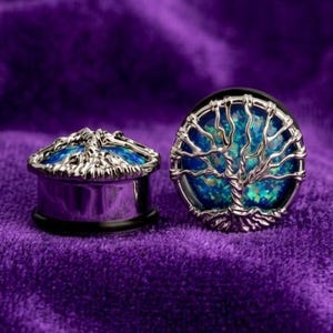 Könnte beinhalten: Zwei silberfarbene Ohrstöpsel mit einem Lebensbaum-Design. Die Stöpsel haben eine blau-grüne, schillernde Mitte. Ein Stöpsel liegt auf der Seite, der andere steht aufrecht. Der Hintergrund ist ein tiefvioletter Samt.