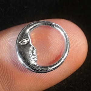 May include: A small, silver-colored crescent moon charm with a detailed face design. The charm is circular with a hollow center and is held on a finger. The moon's face has a closed eye and a defined nose and mouth.