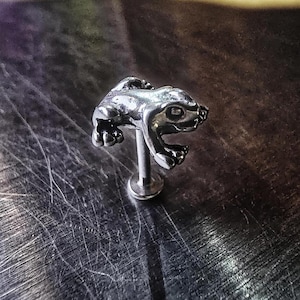 May include: A silver-colored frog-shaped body jewelry stud. The frog is detailed with open mouth and textured skin. The post is a straight, slender rod with a flat, circular base. The jewelry is set against a textured, metallic surface.