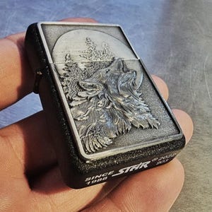 May include: A black and silver lighter featuring a detailed relief of a howling wolf against a moonlit forest scene. The lighter has the text "SINCE 1988 STAR" and the numbers "2022 903" on the bottom.