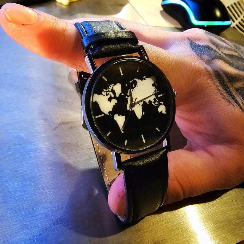 Puede incluir: Un reloj de pulsera de cuero negro con una esfera plateada y un dise&ntilde;o de mapa del mundo blanco. El reloj est&aacute; en la mu&ntilde;eca de una persona.