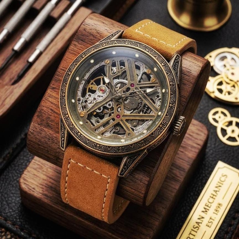 May include: A skeleton watch with a brown leather strap. The watch face showcases intricate mechanical parts in gold and silver hues. The watch is presented on a wooden stand. The text "ARTISAN MECHANICS EST. 1898" is visible.