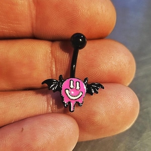 Könnte beinhalten: Ein schwarzer Bauchnabelpiercing mit einem rosa, lächelnden Gesichtsdesign und schwarzen Fledermausflügeln. Das Gesicht hat einen tropfenden Effekt. Die oberen und unteren Kugeln sind schwarz.